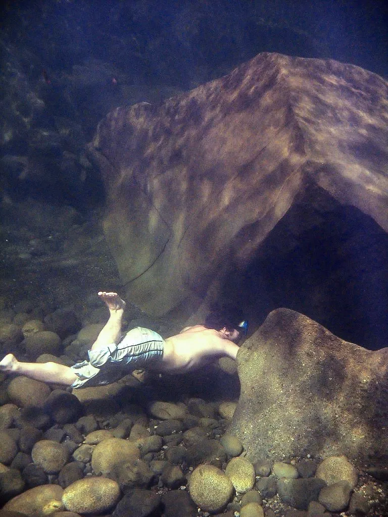 Restauração FiveMeters de Nadadora sob uma saliência de pedras: Pele e shorts azul-brancos separam-se de um leito de água doce verde com pedras cinza e ocres.